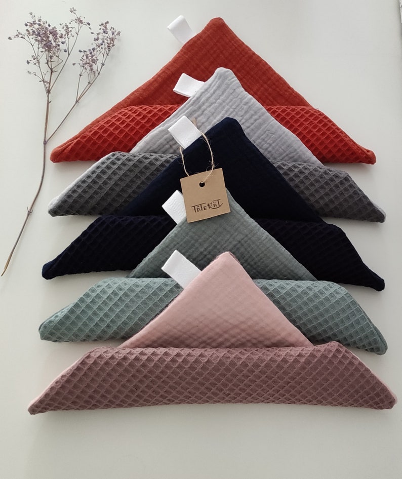 Puede incluir: Un conjunto de siete cuadrados de tejido de tejido de waffle en varios colores, incluyendo rojo, gris, azul marino, verde y rosa. Los cuadrados est&aacute;n doblados en tri&aacute;ngulos y apilados uno encima del otro. Una peque&ntilde;a etiqueta marr&oacute;n con la palabra "Triebel" est&aacute; unida a uno de los cuadrados.