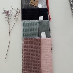 Puede incluir: Una pila de ocho cuadrados de tela de tejido de waffle en varios colores, incluyendo rojo, gris, azul, verde, rosa y marr&oacute;n. Los cuadrados de tela est&aacute;n dispuestos en una pila vertical, con el cuadrado superior siendo rojo y el cuadrado inferior siendo rosa. Una peque&ntilde;a etiqueta marr&oacute;n con el texto "Triksel" est&aacute; unida a la pila.