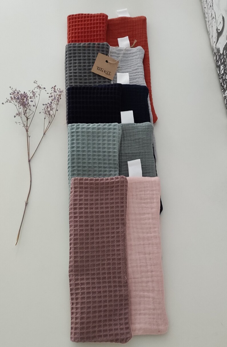 Puede incluir: Una pila de ocho cuadrados de tela de tejido de waffle en varios colores, incluyendo rojo, gris, azul, verde, rosa y marr&oacute;n. Los cuadrados de tela est&aacute;n dispuestos en una pila vertical, con el cuadrado superior siendo rojo y el cuadrado inferior siendo rosa. Una peque&ntilde;a etiqueta marr&oacute;n con el texto "Triksel" est&aacute; unida a la pila.