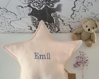 May include: A peach-colored star-shaped pillow with the name "Emil" embroidered in blue thread. The pillow is on a white surface with a small bouquet of purple flowers and a brown tag with the word "Tivoli" on it.