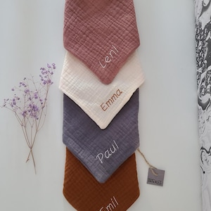 Könnte beinhalten: Vier personalisierte Baby-Lätzchen in verschiedenen Farben: rosa, weiß, grau und braun. Die Lätzchen sind aus weichem, strukturiertem Stoff gefertigt und haben die Namen "Leni", "Emma", "Paul" und "Emil" aufgestickt. Die Lätzchen sind übereinander gestapelt, wobei das rosa Lätzchen oben und das braune Lätzchen unten liegt. Ein kleines braunes Etikett mit dem Wort "INKEL" ist in der unteren rechten Ecke des Bildes sichtbar.