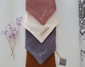 May include: Four personalized baby bibs in different colors: pink, white, gray, and brown. The bibs are made of soft, textured fabric and have the names "Leni", "Emma", "Paul", and "Emil" embroidered on them. The bibs are stacked on top of each other, with the pink bib on top and the brown bib on the bottom. A small brown tag with the word "INKEL" is visible in the lower right corner of the image.