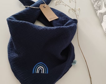 May include: A navy blue bandana with a white and blue rainbow embroidered on it. The bandana is folded in a triangle shape and has a brown tag with the text "The Real" on it.