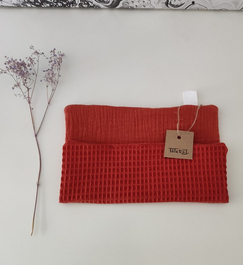 Puede incluir: Una toalla de cocina de tejido de waffle rojo con una capa superior de muselina roja lisa. La toalla tiene una etiqueta marr&oacute;n con el texto "Tater Tot" en ella.