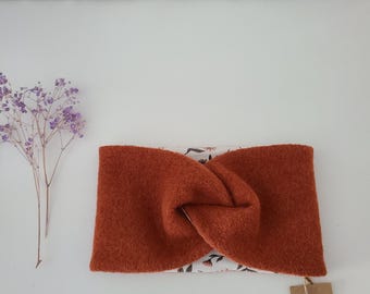 May include: A rust-colored, twisted headband with a floral patterned interior. The headband is made of a soft, textured fabric. A small tag with the text "TaleKat" is attached. A sprig of purple flowers is to the left.