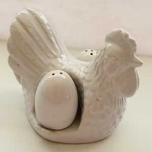 May include: White ceramic rooster-shaped salt and pepper shaker set. The rooster holds two egg-shaped shakers, each with holes for dispensing. The set is a decorative kitchen item.