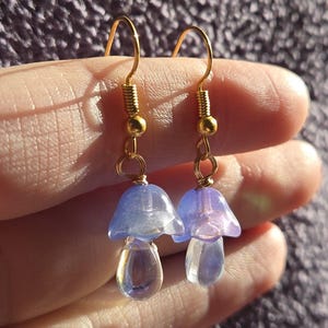 May include: A pair of mushroom-shaped earrings. The earrings have translucent blue caps and clear, teardrop-shaped stems. The ear wires and connecting wires are gold-coloured. The earrings are displayed against a soft, purple background.