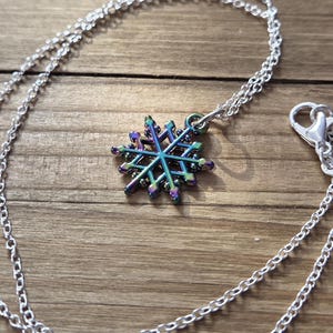 May include: A silver necklace with a snowflake pendant. The pendant is a multi-colored iridescent design with shades of blue, green, and purple. The necklace chain is a delicate link style with a lobster clasp.