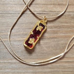 May include: A gold chain necklace with a red and gold pendant. The pendant is shaped like a scroll with gold leaf accents.