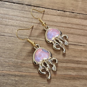 Puede incluir: Un par de pendientes dorados con forma de medusa. Cada pendiente presenta un diseño de medusa iridiscente rosa y blanco con zarcillos colgantes y pequeños cristales transparentes. Los pendientes tienen cierres de gancho de pez dorados.