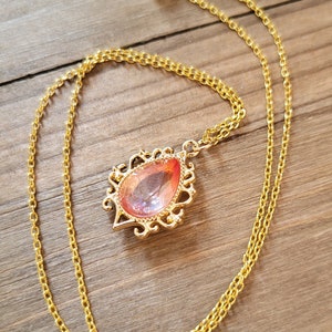 May include: A gold chain necklace with a teardrop-shaped pendant featuring a pink and orange gem set in a gold filigree frame.