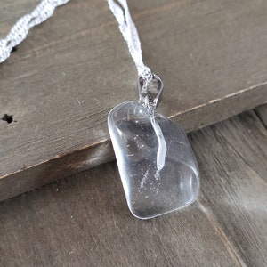 Puede incluir: Un colgante de cristal de cuarzo transparente en una cadena de plata. El colgante tiene forma rectangular con esquinas redondeadas y tiene un ligero aspecto nublado.