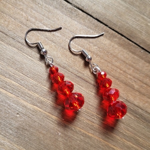 May include: A pair of red beaded earrings with silver-colored fish hook ear wires. The earrings feature a cascading design of faceted red beads, creating a sparkling and eye-catching accessory. The earrings are displayed on a wooden surface.