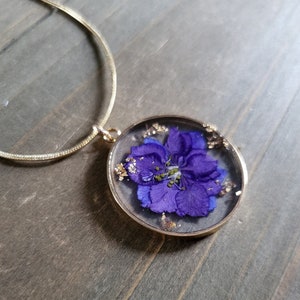 May include: A gold-colored necklace with a round pendant. The pendant contains a vibrant purple flower preserved in clear resin, with gold flakes around the edges. The necklace is displayed on a wooden surface.