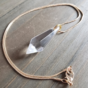 Puede incluir: Un collar de cadena dorada con un colgante de cristal transparente en forma de punta. El colgante está facetado y tiene un aro dorado.