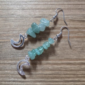 May include: A pair of dangle earrings featuring a silver crescent moon charm and a strand of light green, irregular-shaped beads. The earrings have silver-colored ear wires and are displayed on a wooden surface.