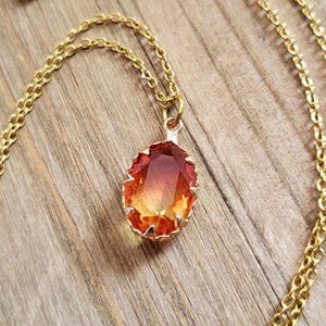 Puede incluir: Un collar de cadena dorada con un gran colgante de forma ovalada con una piedra preciosa roja y naranja.