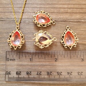 May include: Four gold-tone teardrop-shaped pendants with clear pink rhinestones. The pendants are approximately 1 inch in length and are shown next to a ruler.