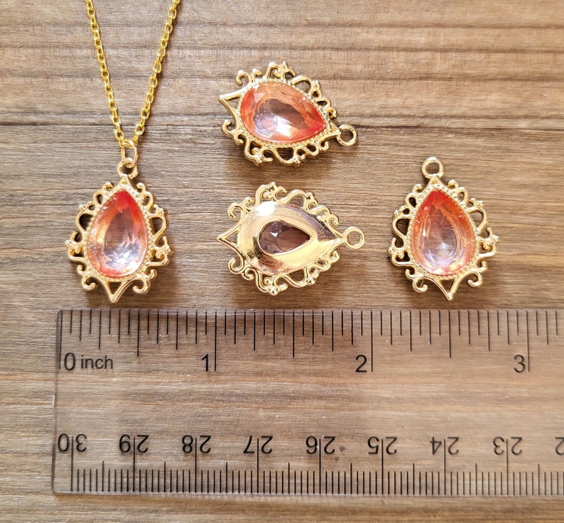 May include: Four gold-tone teardrop-shaped pendants with clear pink rhinestones. The pendants are approximately 1 inch in length and are shown next to a ruler.