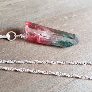 May include: A silver chain necklace with a pink, green, and black crystal pendant. The pendant is shaped like a long, thin point.