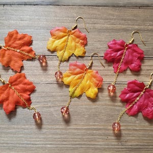 Puede incluir: Cinco pares de pendientes colgantes con dijes de hoja de arce en tonos rojos, naranjas y amarillos. Cada dije de hoja está unido a una cadena dorada con una pequeña cuenta transparente.