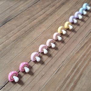 May include: A row of colorful mushroom-shaped embellishments. The mushrooms have pink, yellow, purple, and blue caps with white stems. One mushroom has a pink cap with white polka dots. The mushrooms are arranged diagonally on a wooden surface.