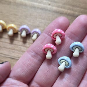 May include: A collection of colorful mushroom-shaped charms. The charms feature a variety of colors, including yellow, pink, blue, and purple. Each mushroom has a white stem and a dotted cap. The charms are small and appear to be made of resin or a similar material.