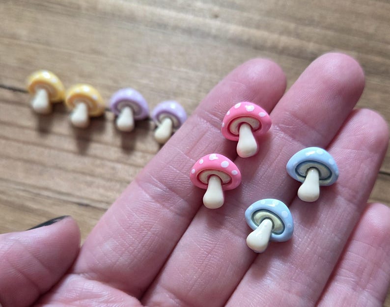 May include: A collection of colorful mushroom-shaped charms. The charms feature a variety of colors, including yellow, pink, blue, and purple. Each mushroom has a white stem and a dotted cap. The charms are small and appear to be made of resin or a similar material.