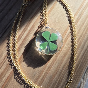 May include: A gold-colored chain necklace with a pendant featuring a four-leaf clover encased in clear resin. The pendant has a gold-colored border. The necklace rests on a wooden surface.