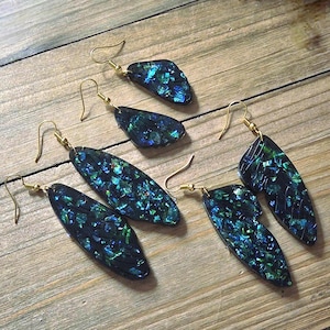 May include: Six pairs of earrings with gold-coloured hooks. The earrings have a dark base with iridescent blue and green accents. The earrings are in various shapes, including teardrop and wing-like designs. The earrings are displayed on a wooden surface.