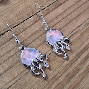May include: A pair of silver-toned jellyfish earrings. Each earring features a pearlescent, pink and white jellyfish design with dangling tentacles and small, clear crystals. The earrings have a hook closure.
