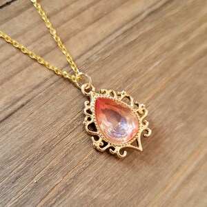 May include: A gold chain necklace with a pendant featuring a large, teardrop-shaped, iridescent pink gemstone set in a gold filigree frame.
