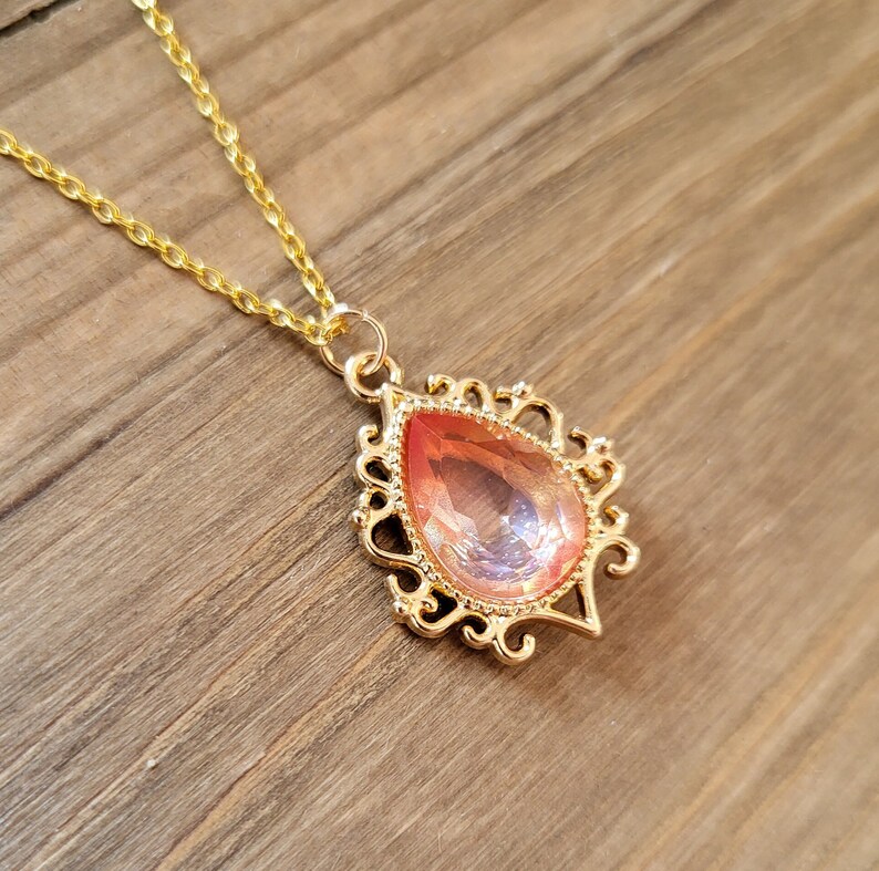 May include: A gold chain necklace with a pendant featuring a large, teardrop-shaped, iridescent pink gemstone set in a gold filigree frame.