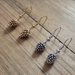 May include: Pine cone earrings in gold and silver tones. The earrings feature a detailed pine cone charm suspended from a hook. The gold earrings have a textured finish, while the silver earrings have a polished look. The earrings are displayed on a wooden surface.