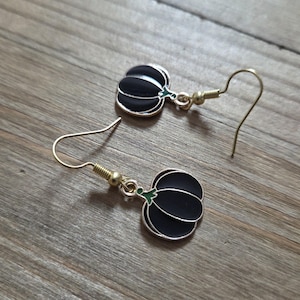 Op de afbeelding: Een paar goudkleurige oorhaken met zwarte pompoenvormige bedels en groene steeltjes. De oorbellen worden getoond op een houten oppervlak, wat hun herfstontwerp benadrukt. Een charmant accessoire.