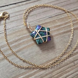 May include: A gold-coloured chain necklace with a star-shaped pendant. The pendant is a mix of green and purple stone, wrapped in gold wire. The necklace is displayed on a wooden surface.