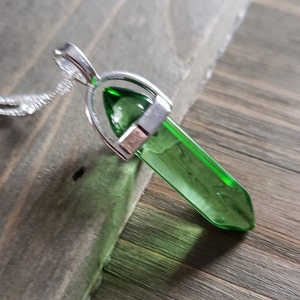 Op de afbeelding: Een zilveren ketting met een groene kristallen hanger. De hanger is puntig en heeft een zilveren setting.