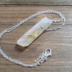 May include: A silver chain necklace with a clear quartz crystal pendant. The crystal is a long, thin point with a natural, rough texture.