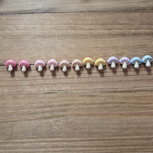 May include: A row of eleven miniature mushroom-shaped embellishments in various pastel colors, including pink, yellow, and blue. Some mushrooms have white polka dots on their caps. Each mushroom has a white stem.