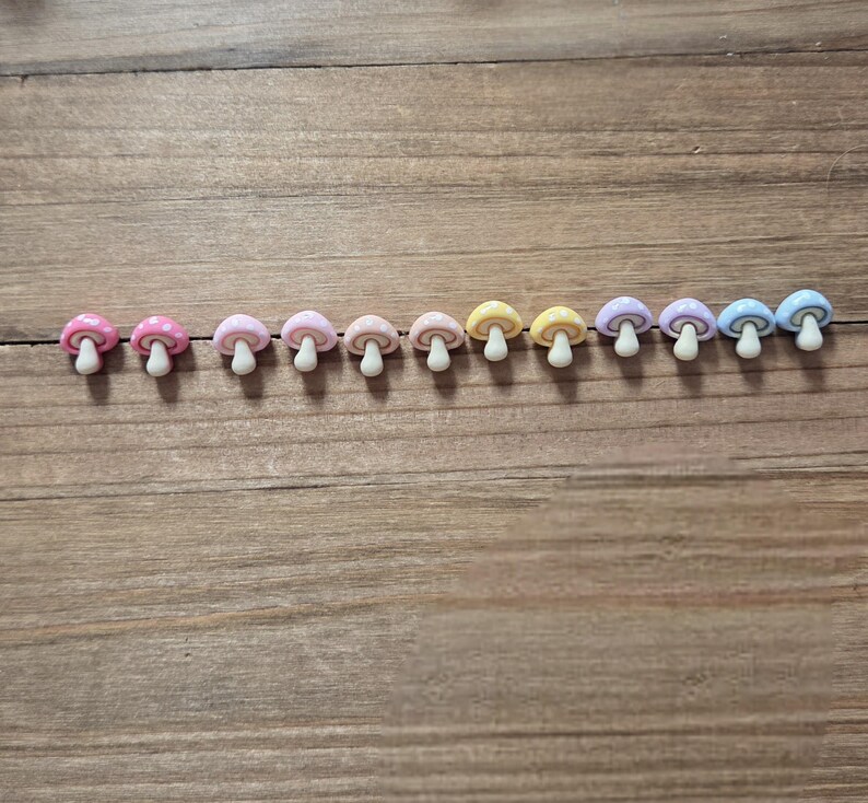 May include: A row of eleven miniature mushroom-shaped embellishments in various pastel colors, including pink, yellow, and blue. Some mushrooms have white polka dots on their caps. Each mushroom has a white stem.