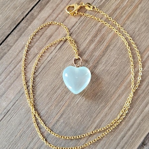 May include: A gold-colored chain necklace with a light blue heart-shaped pendant. The pendant is smooth and translucent. The necklace is displayed on a wooden surface.