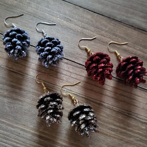 May include: Four pairs of dangle earrings featuring glitter-covered pine cone shapes. The earrings are available in silver, gold, and red.