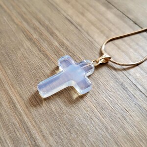 May include: A cross pendant necklace with a gold chain. The cross is made of a translucent, opalescent material, with a soft, glowing appearance. The necklace is displayed on a wooden surface.
