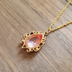 May include: A gold chain necklace with a pink teardrop-shaped pendant. The pendant has a detailed ornate design.