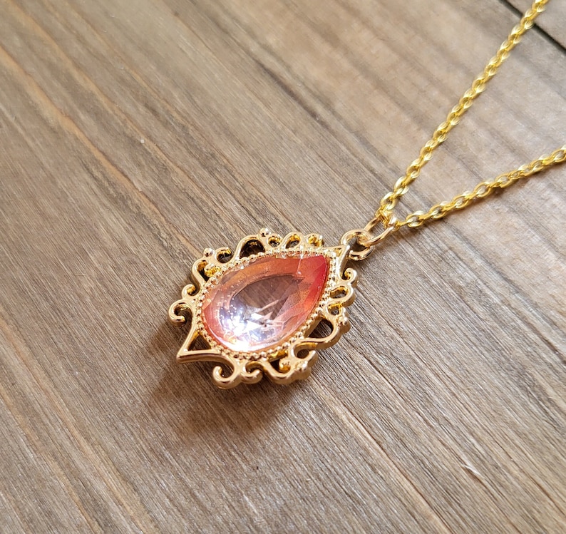 May include: A gold chain necklace with a pink teardrop-shaped pendant. The pendant has a detailed ornate design.