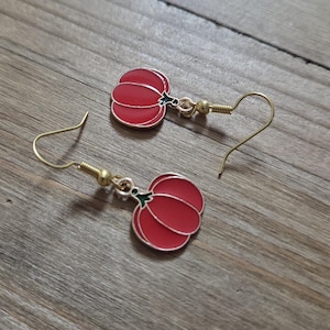 Puede incluir: Un par de pendientes rojos con forma de calabaza con ganchos dorados. Los pendientes tienen un contorno dorado y detalles verdes en el tallo. Se muestran sobre una superficie de madera.