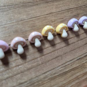 May include: A string of colorful mushroom-shaped beads. The beads are in various pastel colors, including pink, yellow, and purple, with white stems. The mushrooms are strung together on a dark brown string, creating a whimsical and decorative element.