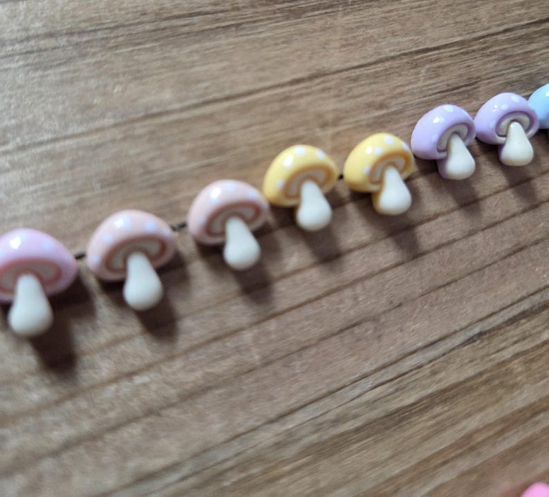May include: A string of colorful mushroom-shaped beads. The beads are in various pastel colors, including pink, yellow, and purple, with white stems. The mushrooms are strung together on a dark brown string, creating a whimsical and decorative element.