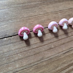 May include: A row of small, pink mushroom-shaped decorative items with white polka dots on the caps and white stems. The mushrooms are arranged on a wooden surface, creating a whimsical and playful aesthetic.