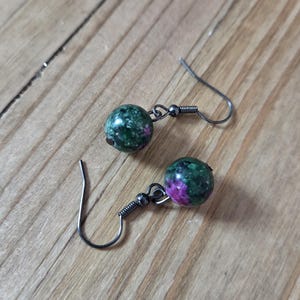 May include: A pair of earrings featuring round, green and purple gemstone beads. The beads are attached to silver-colored fish hook ear wires. The earrings are displayed on a wooden surface.